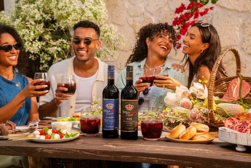 Friends share a sunny brunch with semi-sweet Stella Rosa Blueberry and Stella Rosa Black wines, paired with croissants, bread, cheese, cured meat, tomatoes, basil, grapes, strawberries, watermelon, and other light bites.